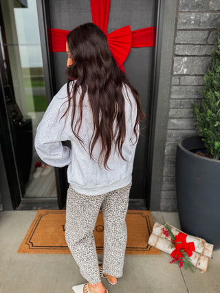 A woman with long dark brown hair stands with her back to the camera, wearing a light gray sweatshirt and leopard-print pants, standing on a doormat in front of a gray door decorated with a large red bow.