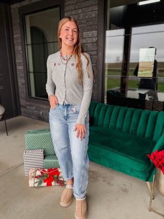 A woman stands on a porch wearing a light gray knit zip-front cardigan with small embroidered details, light-wash blue jeans, and tan ankle boots. A green velvet sofa, wrapped holiday gifts, and a brick exterior are visible in the background.