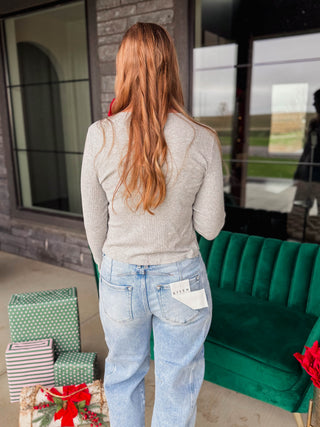 A woman with long hair is shown from behind, wearing a light gray long-sleeve top and light blue jeans with a white tag attached to the back pocket. She is standing on a patio in front of a modern house, with wrapped Christmas gifts on the left and a green velvet sofa on the right.
