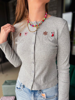 A woman wearing a light gray long-sleeve button-front cardigan with embroidered holiday motifs, paired with blue jeans and layered necklaces.