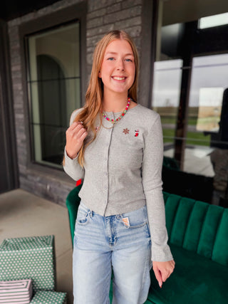 A smiling young woman stands on a porch wearing a light gray long-sleeve button-front cardigan with small holiday-themed embroidered pins, light-wash blue jeans, and layered necklaces. A green velvet sofa and wrapped gift boxes are visible in the background.
