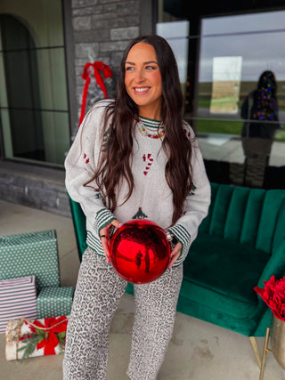 A woman wearing a cozy Christmas-themed knit sweater and matching patterned pants stands in a decorated home setting, holding a large red ornament. The sweater is off-white with green striped trim and red candy cane motifs, paired with matching off-white pants featuring a gray and white pattern.