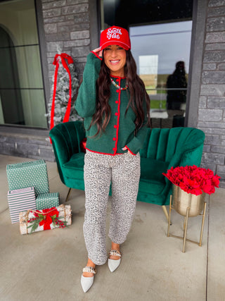 A woman stands on a patio wearing a festive holiday outfit consisting of a green knit cardigan with red buttons, matching green and red patterned pants, a red baseball cap with white text, and white pointed flats. Behind her is a green velvet sofa, wrapped gift boxes, and red flowers.