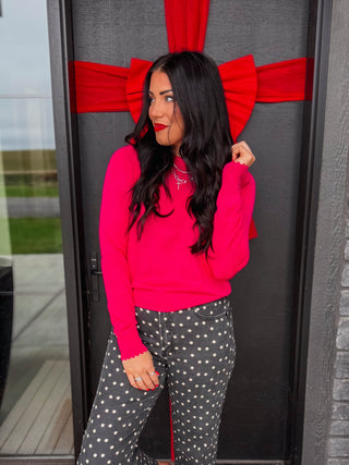 A woman stands on a welcome doormat in front of a dark gray door decorated with a large red bow. She is wearing a bright hot pink long-sleeve top, dark gray wide-leg pants with white polka dots, and white pointed-toe flats.