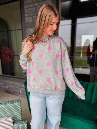 A woman wearing a light gray crewneck sweatshirt with bright pink polka dots, paired with light blue jeans and a chunky gold necklace, standing in front of a modern glass storefront.