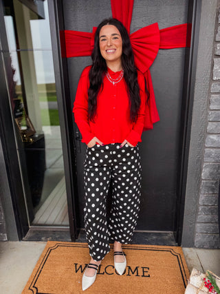 A woman stands in front of a dark gray door decorated with a large red bow, wearing a bright red long-sleeve button-up shirt, black wide-leg pants with white polka dots, and white pointed-toe flats on a brown doormat.