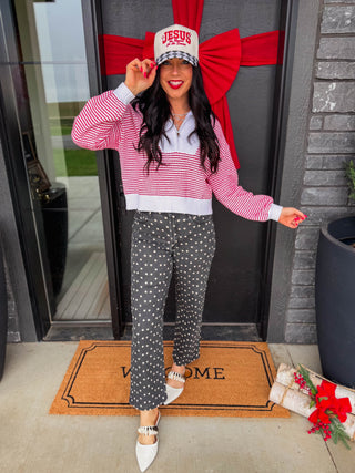 A woman stands in front of a house door wearing a pink and white striped long-sleeve crop top, dark gray pants with white polka dots, white pointed flats, and a white baseball cap with red text that reads 'JESUS IS REASON FOR THE SEASON '. A large red bow decorates the door behind her