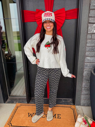 A woman stands in front of a decorated front door wearing a cream long-sleeve crewneck sweater with a red and green Christmas graphic, dark gray pants with white polka dots, and a white cap that says 'JESUS IS THE REASON FOR THE SEASON'.