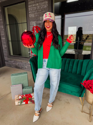 A woman stands on a patio wearing a festive holiday outfit with a green blazer, red top, light blue jeans, white pointed shoes, and a beige cap that reads 'MERRY & BRIGHT'. She holds a large red ornament next to a green velvet sofa and wrapped gift boxes.