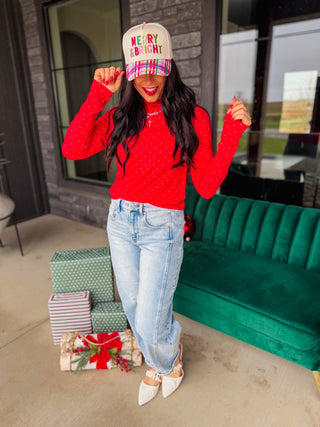 A woman posing on a patio wearing a red long-sleeve knit top with white polka dots, light-wash jeans, white pointed-toe heels, and a beige trucker hat that reads 'MERRY & BRIGHT' in colorful letters. A green velvet sofa and wrapped holiday gifts are visible in the background.