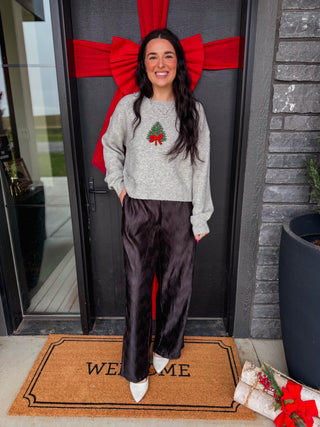 A woman stands on a front porch wearing a light gray crewneck sweatshirt with a green and red Christmas tree graphic, paired with black wide-leg satin pants and white pointed shoes. A brown 'WELCOME' doormat and a large red bow on a black door are visible behind her.