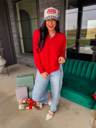 A woman stands on a porch wearing a bright red long-sleeve sweater, light blue jeans, white pointed flats, and a white baseball cap with red 'JESUS' lettering. A green velvet sofa, wrapped gifts, and large windows are visible in the background.