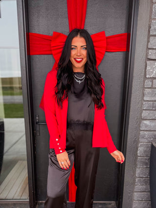 A woman stands in front of a house entrance wearing a red blazer, black satin jumpsuit, and white pointed flats. A large red bow is tied behind her on a dark door, with a welcome doormat and holiday decorations nearby.