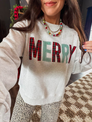 A woman wearing a light heather gray sweatshirt with the word 'MERRY' in large red, green, and black plaid letters, paired with leopard-print pants and layered necklaces.