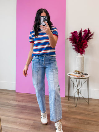 Person taking a mirror selfie wearing a striped shirt and jeans in front of a pink wall.