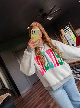 A woman takes a mirror selfie wearing a white crewneck sweatshirt with a colorful 'JOLLY' graphic in pink, red, and green, paired with light blue jeans in a dark room.